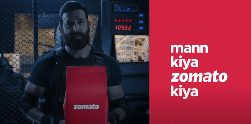 Screenshot einer Werbe-Kampagne von Zomato: Auf der linken Seite hält der indische Schauspieler Hrithik Roshan eine rote Zomato-Papiertüte in die Kamera; die rechte Hälfte zeigt den Slogan „mann kiya zomato kiya“ in weißer Schrift auf rotem Hintergrund.