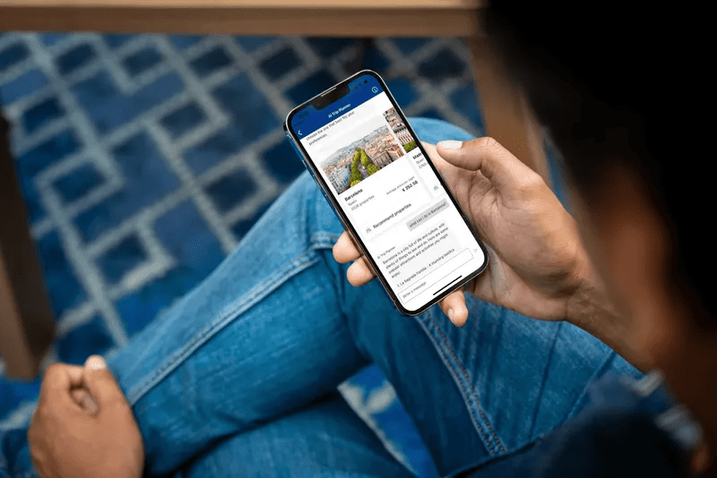 Eine Person hält ein Smartphone in der Hand, auf dem die Benutzeroberfläche eines mobilen „AI Trip Planner“ mit Reisevorschlägen und Preisangaben für Barcelona geöffnet ist.