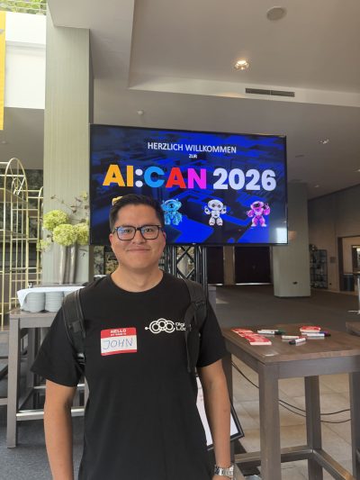 John Munoz standing in front of a screen showing "AI:CAN 2026"